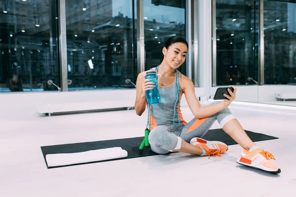 Sorrindo Asiático Esportista Sentado Tapete Fitness Segurando Esportes Garrafa Com — Fotografia de Stock