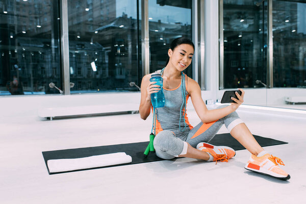 smiling asian sportswoman sitting on fitness mat, holding sports bottle with water and taking selfie at gym