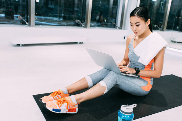 attractive asian girl with towel on shoulders sitting on fitness mat and typing on laptop keyboard at gym