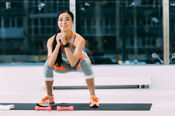 smiling asian girl doing squats on fitness mat at gym