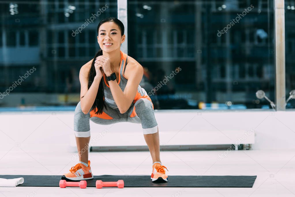 smiling asian girl doing squats on fitness mat at gym