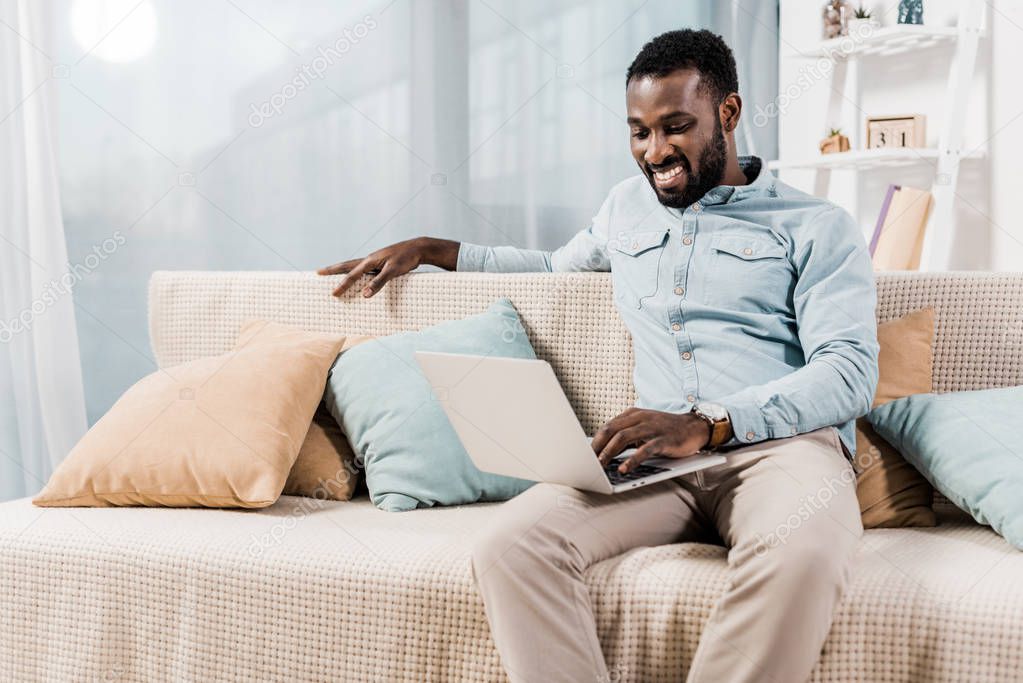 african american freelancer sitting on couch in living room and using laptop