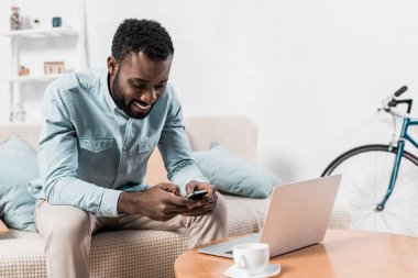 african american freelancer sitting on couch and typing on smartphone clipart