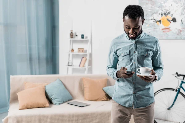 Улыбающийся Африканский Американец Печатающий Смартфоне Держащий Чашку Кофе — Бесплатное стоковое фото