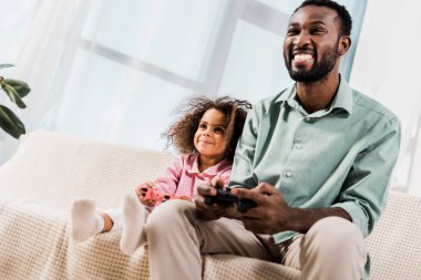 african american man and daughter playing with play stations in living room clipart