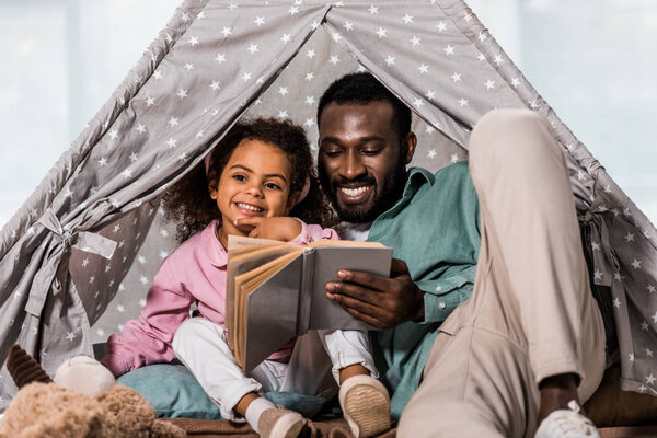 african american father reading book with child and smiling in living room