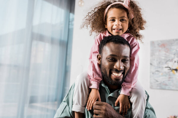 african american dad carrying daughter on neck in living room