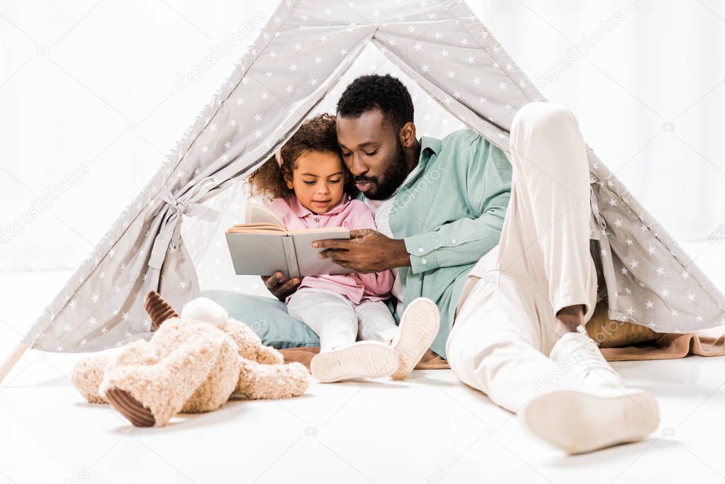 african american man and daughter reading book in living room