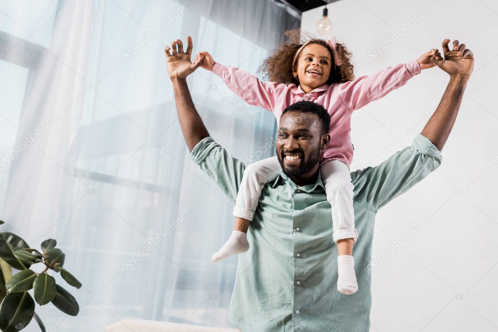 african american father carrying daughter on neck and playing in living room