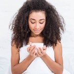 Curly african american girl holding cup of coffee in bed during morning time at home