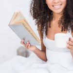 Partial view of african american girl holding coffee cup and reading book in bed during morning time at home