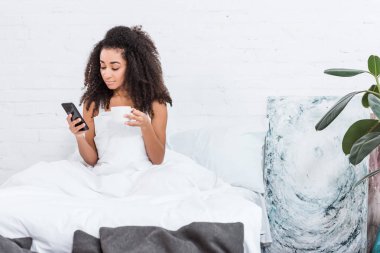 Afrikalı-Amerikalı genç kadın ile kahve fincan smartphone yatakta evde sabah zaman sırasında kullanarak