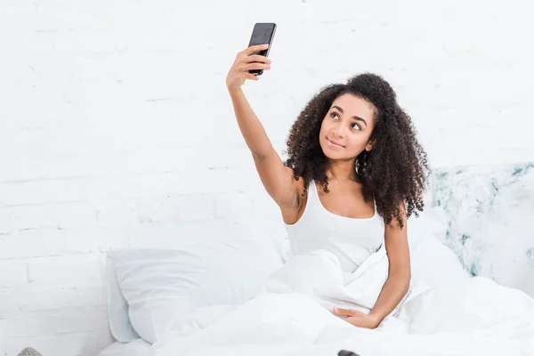 有吸引力的非洲裔美国女孩在早上在家的时候在床上的智能手机自拍自拍 — 图库照片