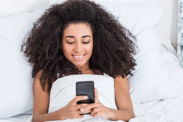 快乐的非洲裔美国年轻女子在早上在家中使用智能手机在床上 — 免费的图库照片