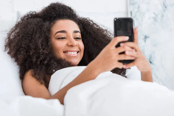 选择重点的非洲美国年轻女子使用智能手机在床上上午在家里的时间 — 免费的图库照片