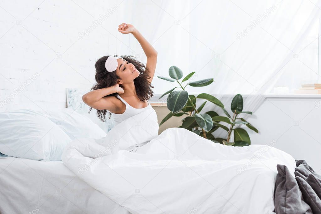 side view of young african american woman with blindfold on forehead stretching in bed during morning time at home