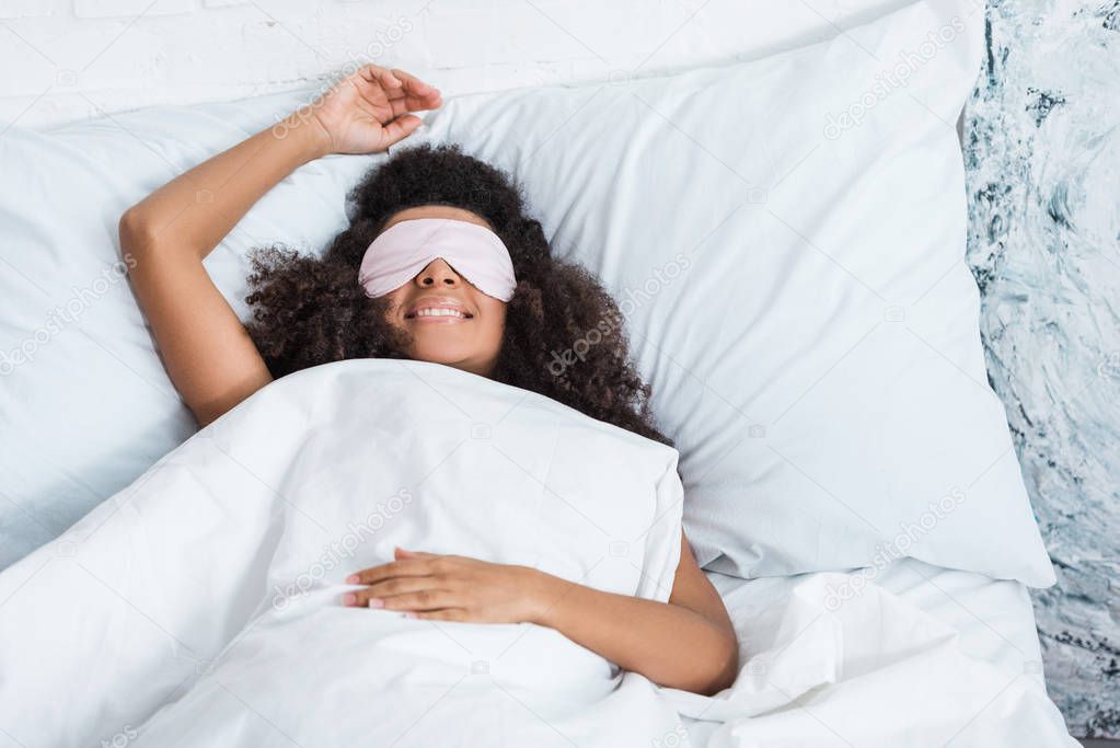 smiling african american girl with eyes covered by sleeping blindfold in bed during morning time at home