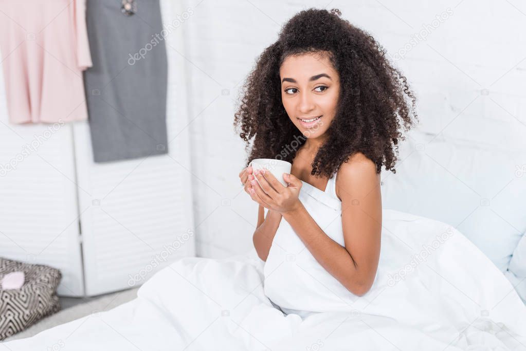 beautiful curly african american girl holding cup of coffee in bed during morning time at home