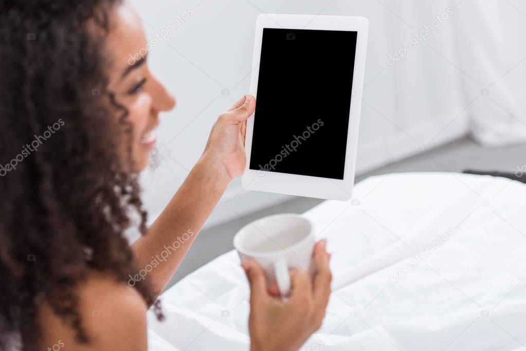 selective focus of african american woman with coffee cup using digital tablet in bed at home
