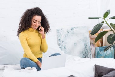 smartphone üzerinde konuşmak ve evde yatakta dizüstü bilgisayar üzerinde çalışan kıvırcık Afrikalı-Amerikalı kadın serbest meslek