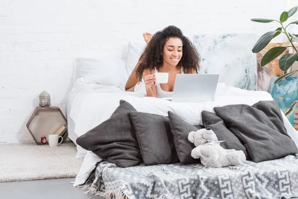 有吸引力的快乐女孩拿着咖啡杯 并在床上使用笔记本电脑在早上的时间在家里 — 图库照片