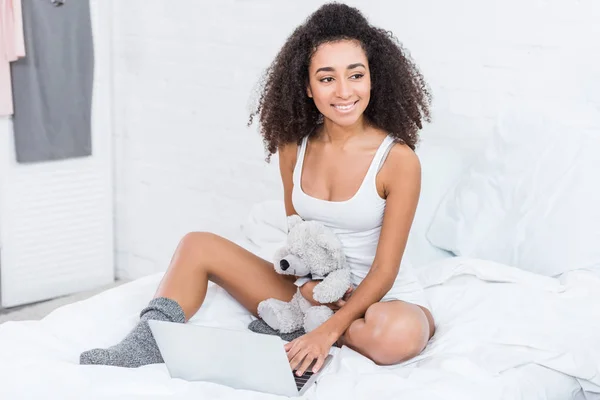 Σγουρά Νεαρή Γυναίκα Κρατώντας Αρκουδάκι Και Χρησιμοποιώντας Laptop Στο Κρεβάτι — Δωρεάν Φωτογραφία