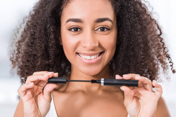 cheerful curly african american girl using mascara and looking at camera