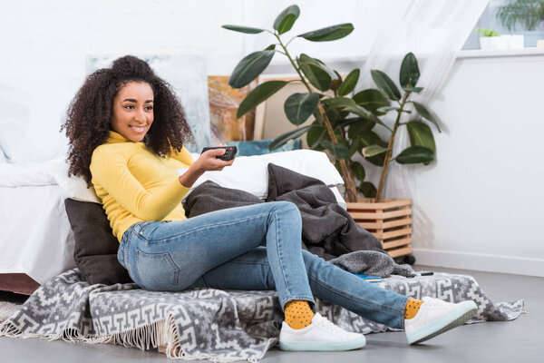 stylish african american girl using remote controller and watching tv at home