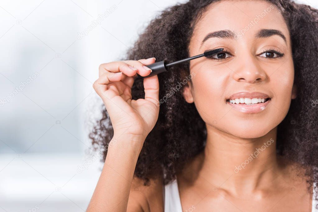 close up view of curly african american girl using mascara and looking at camera