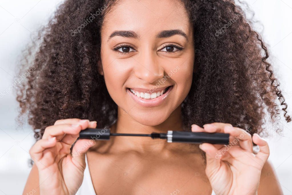 cheerful curly african american girl using mascara and looking at camera