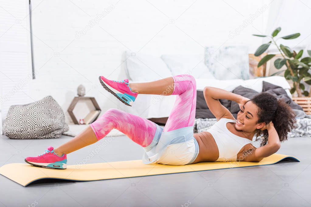smiling african american sportswoman doing abs on fitness mat in bedroom at home