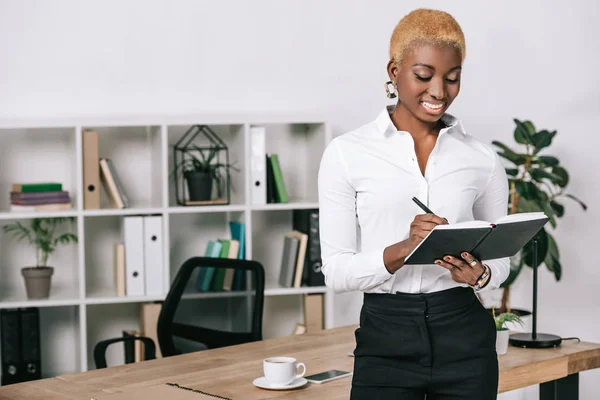 Щаслива Афроамериканська Бізнес Леді Коротким Волоссям Пише Блокноті Сучасному Офісі — Безкоштовне стокове фото