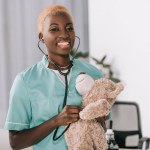 Sonriente africana americana enfermera examinando osito de peluche con estetoscopio