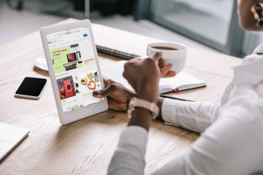 Kırpılan fincan kahve ve dijital tablet ile ebay app ekran üzerinde tutan Afrikalı-Amerikalı kadın görünümünü