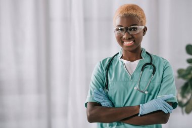 smiling african american nurse in gloves standing with crossed arms while looking at camera clipart