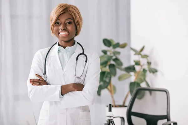 聴診器立って両手を交差しながらカメラ目線で魅力的な笑顔のアフリカ系アメリカ人女医 — ストック写真