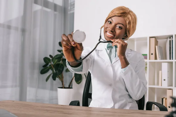 Lächelnder Afrikanisch Amerikanischer Arzt Zeigt Stethoskop Während Der Klinik Sitzt — kostenloses Stockfoto