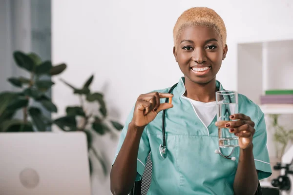 錠剤と水のガラスを保持している聴診器でアフリカ系アメリカ人看護師笑顔  — 無料ストックフォト