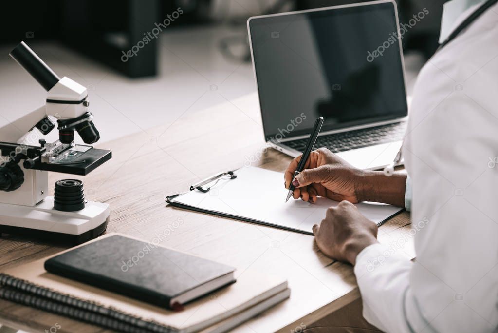 cropped view of african american scientist writing on paper near laptop with blank screen 