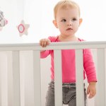 Adorable child in pink shirt standing in crib and looking away
