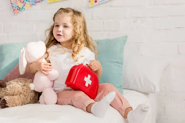 adorable kid holding first aid kit and rabbit toy in children room, looking away