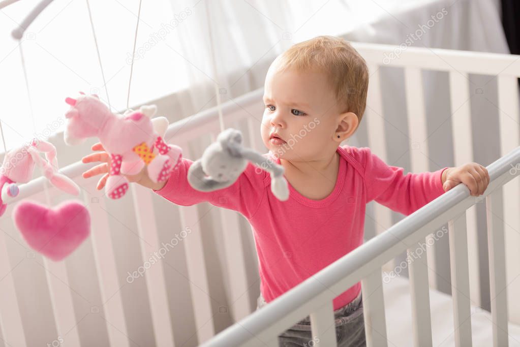 adorable child in pink shirt standing in crib and touching toys