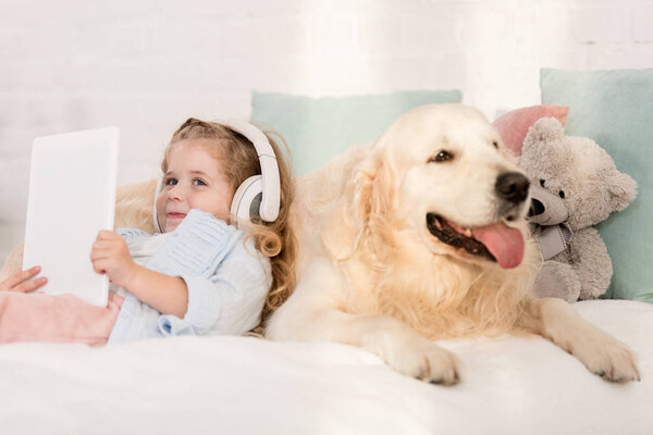 adorable kid listening music with tablet and leaning on golden retriever on bed in children room