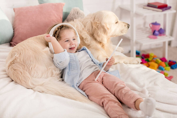 adorable kid using tablet and leaning on golden retriever on bed in children room