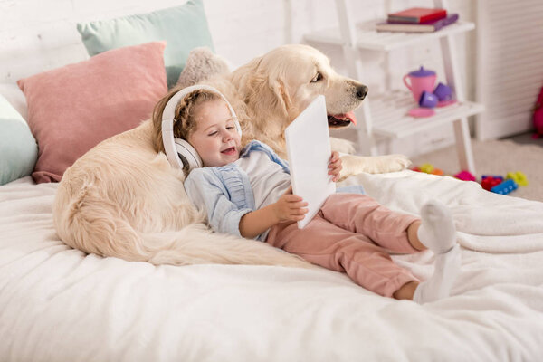 adorable happy kid listening music with tablet and leaning on golden retriever dog on bed in children room