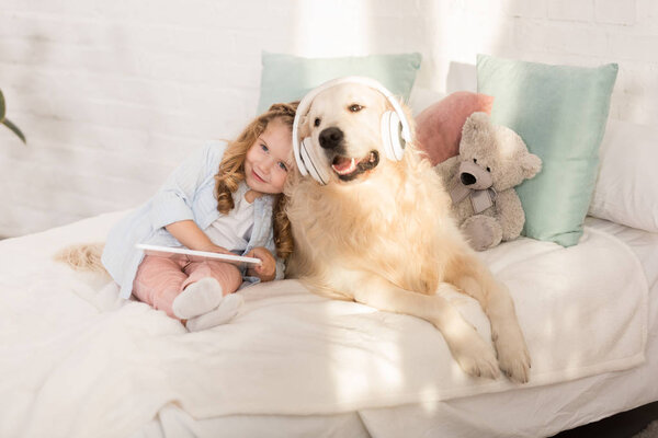 adorable kid holding tablet and leaning on golden retriever with headphones lying on bed in children room