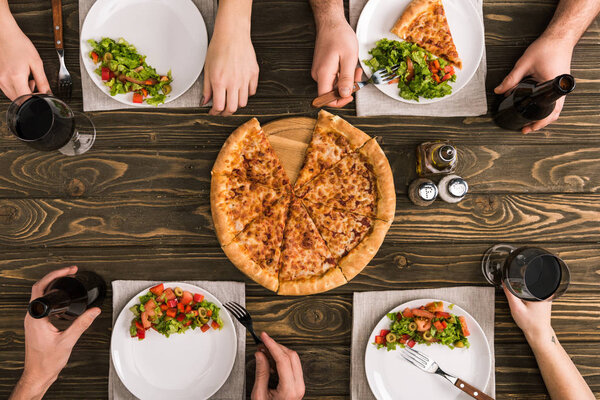 частичный вид друзей, обедающих с пиццей и салатами за деревянным столом
