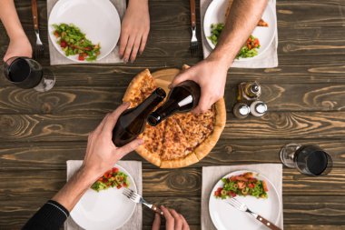 cropped view of friends toasting while having dinner with pizza and salads at wooden table clipart