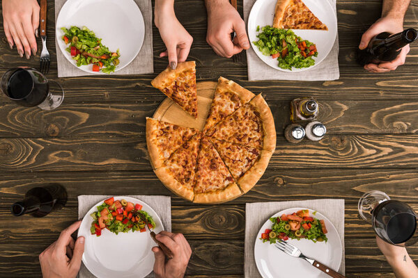 частичный вид друзей, обедающих с салатами и пиццей за деревянным столом
