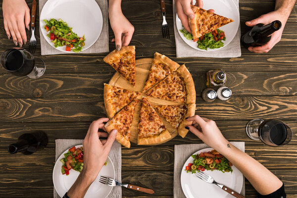 частичный вид друзей, делящих пиццу за ужином с салатами за деревянным столом
